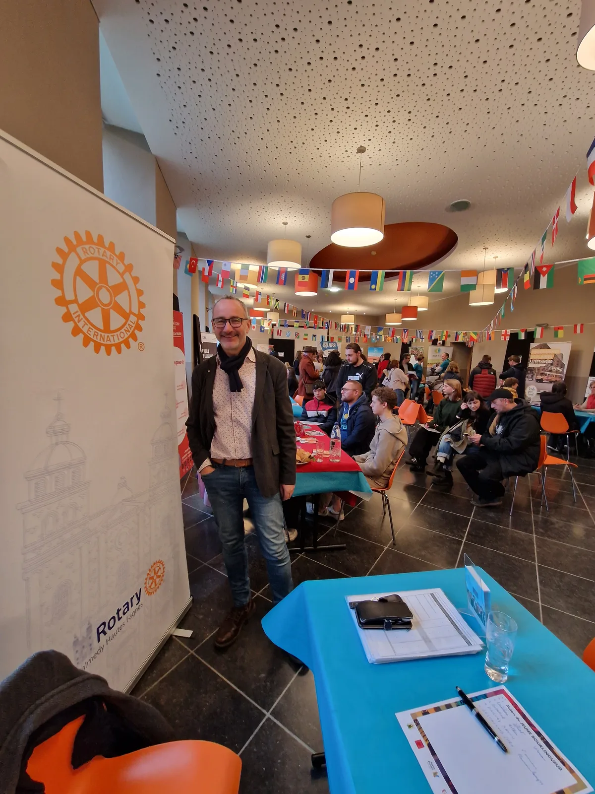 Un membre du club devant le stand du Rotary Malmedy Hautes-Fagnes, dans une salle décorée de drapeaux du monde entier où des jeunes s'informent